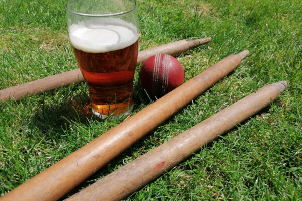 A pint sits on the grass inbetween some cricket stumps and a cricket ball