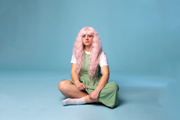Millie has pink hair and is wearing a green dress with a white t-shirt underneath. She is sitting cross-legged on the floor with a blue backdrop.